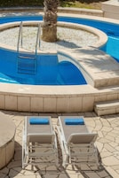 A swimming pool with a palm tree, two lounge chairs, and a stone-tiled deck.