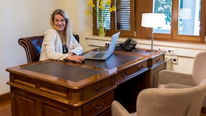 A person seated at a wooden desk with a laptop, a telephone, and a vase with yellow flowers.