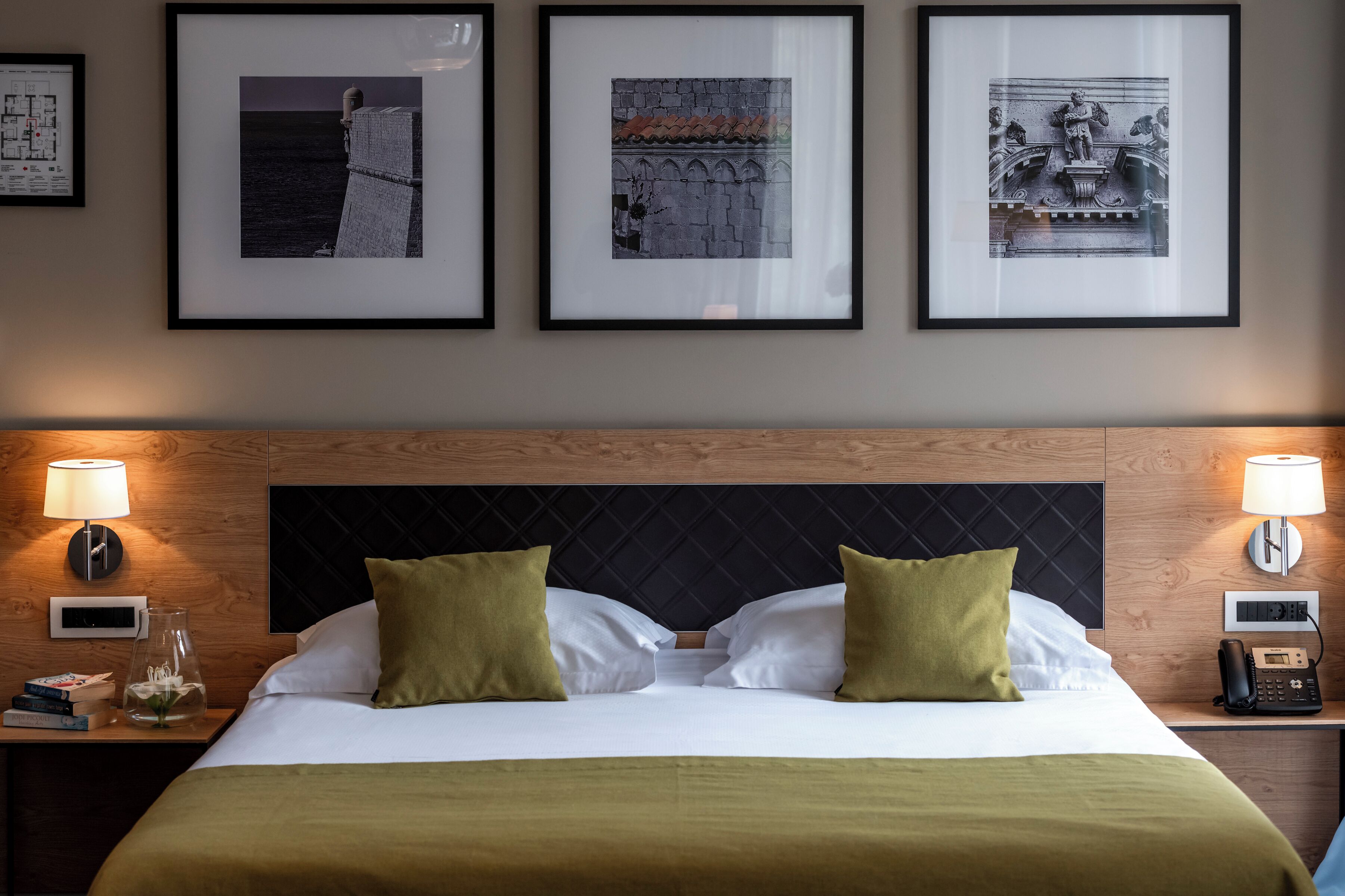 Habitación de hotel con una cama, dos lámparas de noche, un mesita de noche con un teléfono y tres cuadros en la pared.