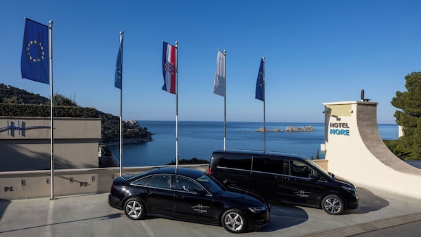 A black car parked in front of a building with flags, including the European Union flag, and a view of the sea.