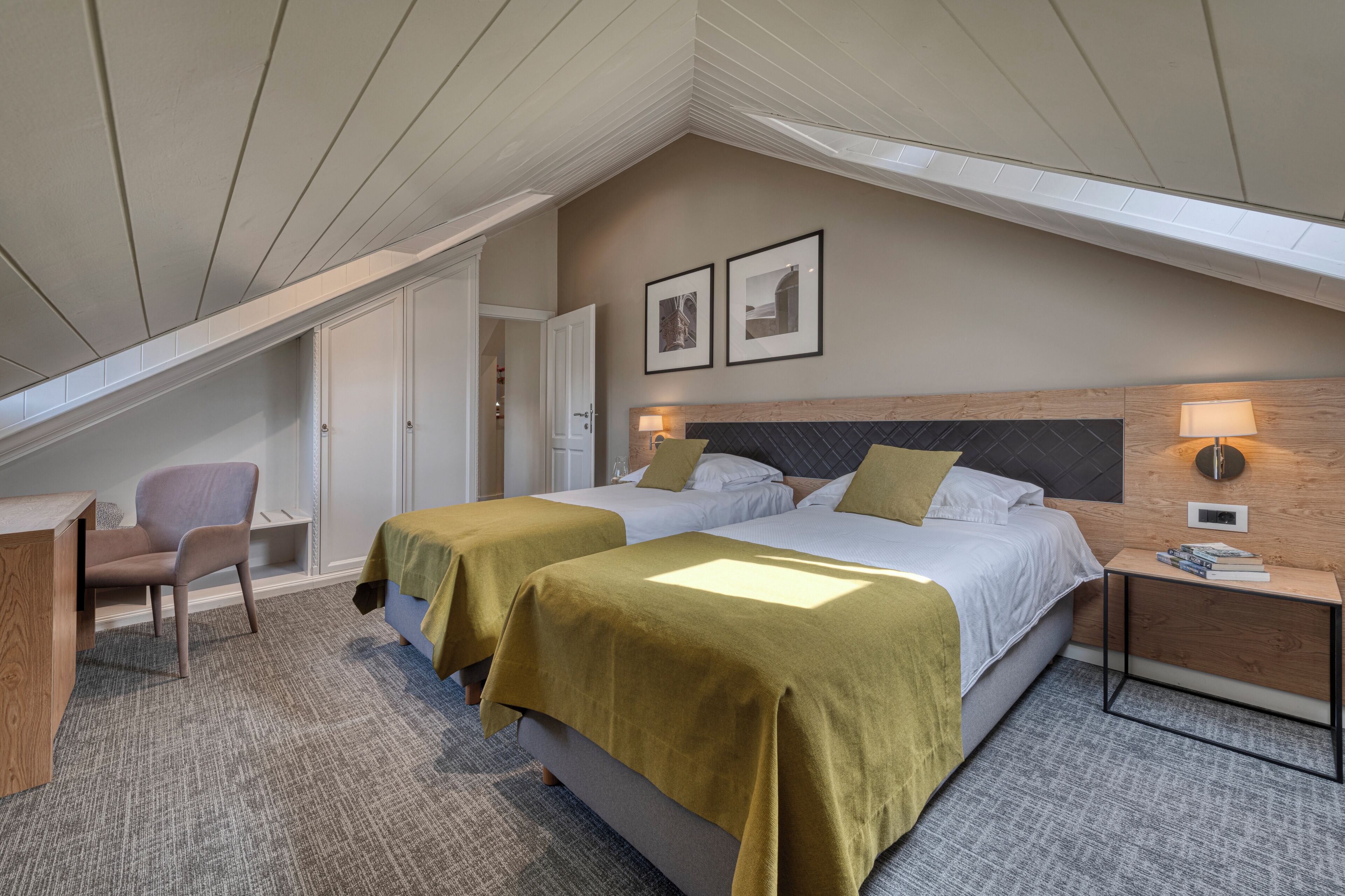 Quarto de hotel com duas camas, uma escrivaninha, uma cadeira e uma pequena mesa com abajur.