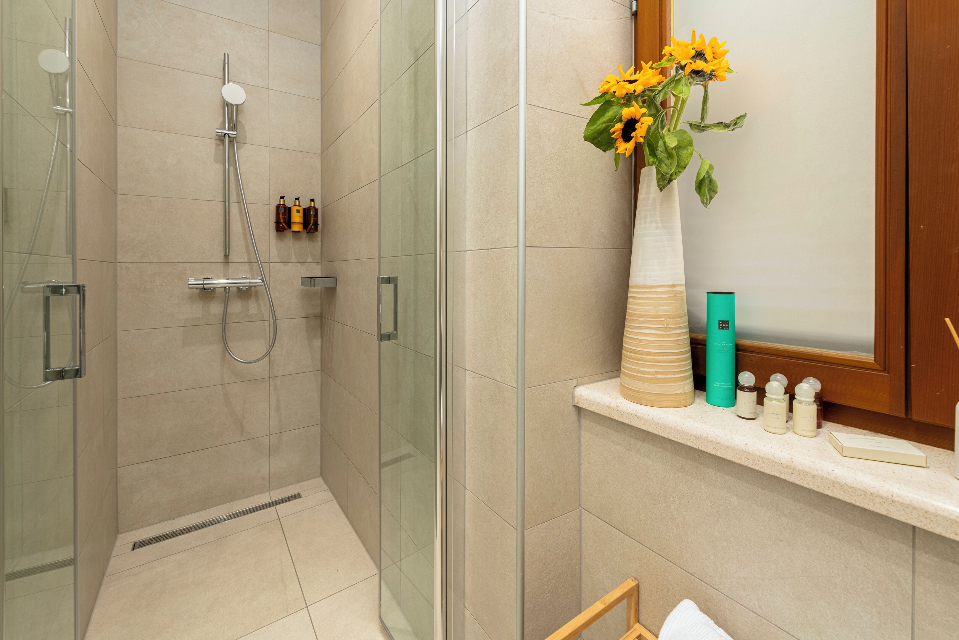 Un baño con ducha de vidrio, un estante con artículos de tocador y un jarrón con flores.