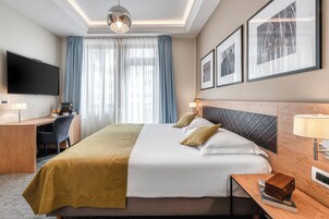 Quarto de hotel com cama, escrivaninha, televisão e janela com cortinas.