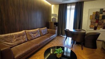 A modern living room with a brown leather sofa, a round black coffee table with a plant, a wooden floor, a dining table with chairs, and a wall art piece.