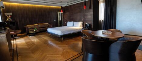 A modern hotel room with a wooden floor, a bed, a desk, and a dining area with chairs.