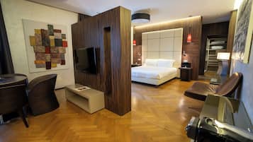 A modern hotel room with a wooden floor, a large bed, a TV, a desk, and a seating area.