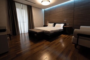 A modern hotel room with a wooden floor, a bed with white linens, a small bedside table, and a chair.