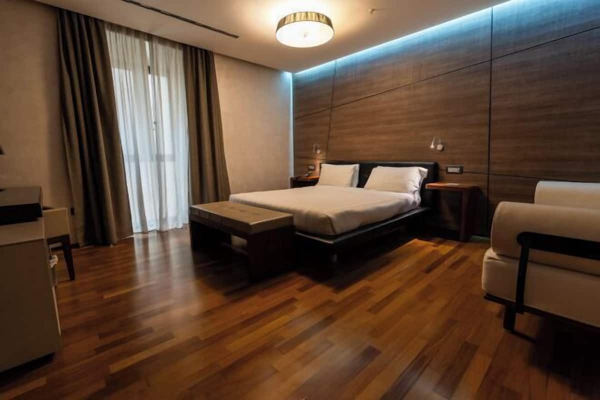 A modern hotel room with a wooden floor, a bed with white linens, a bedside table, a lamp, and a chair.