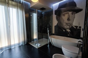 A modern bathroom with a glass-enclosed shower, a freestanding bathtub, and a large mirror with a portrait.