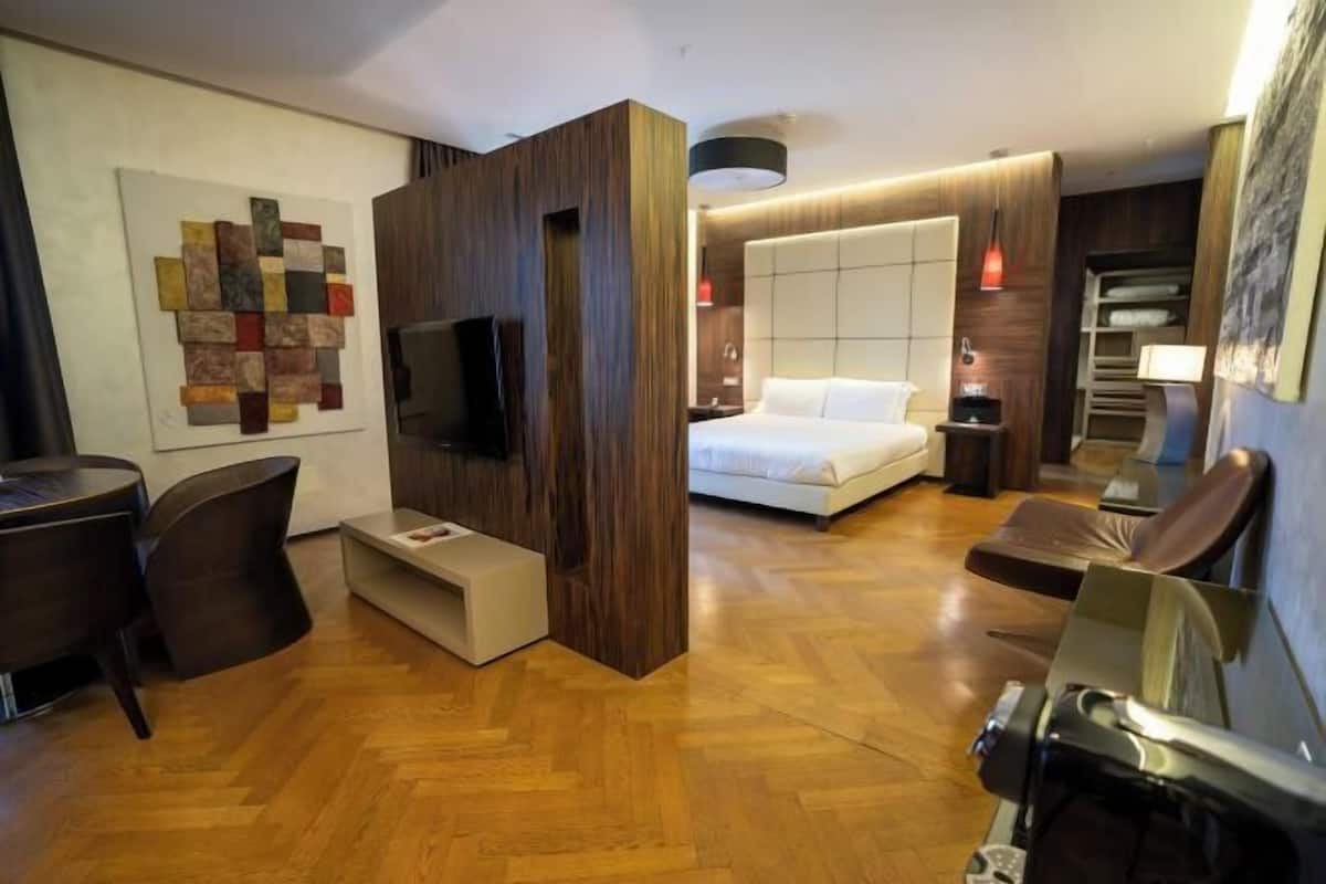 A modern hotel room with a wooden floor, a bed with a headboard, a TV mounted on a wooden panel, a desk with chairs, and a wall art piece.
