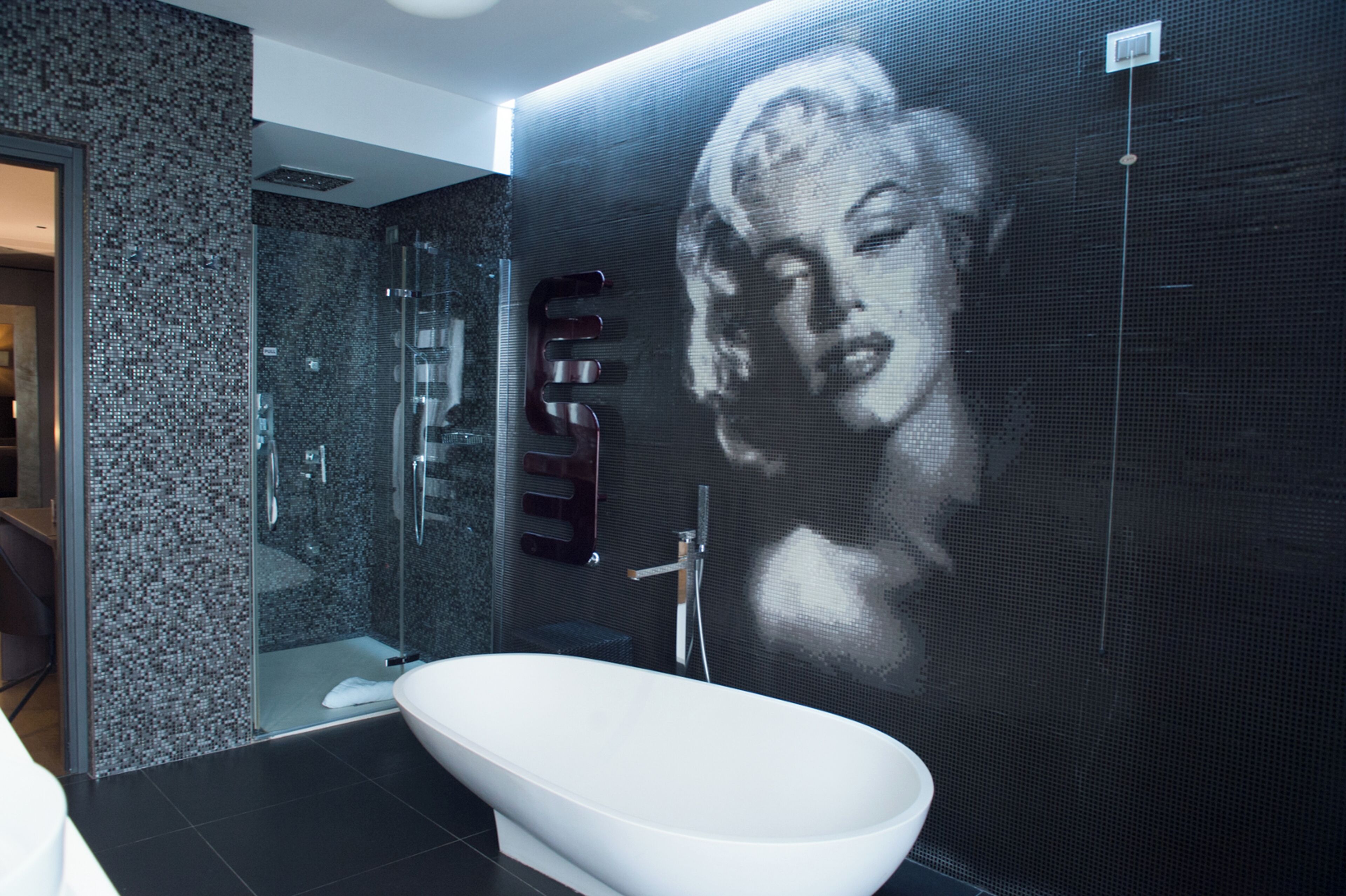 A modern bathroom with a large digital screen displaying a portrait, a glass-enclosed shower, and a white bathtub.