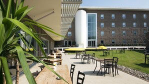 An outdoor patio area with tables and chairs, a large building in the background, and a grassy lawn. - Aiden by Best Western JHD Dunant Hotel (Castiglione delle Stiviere)