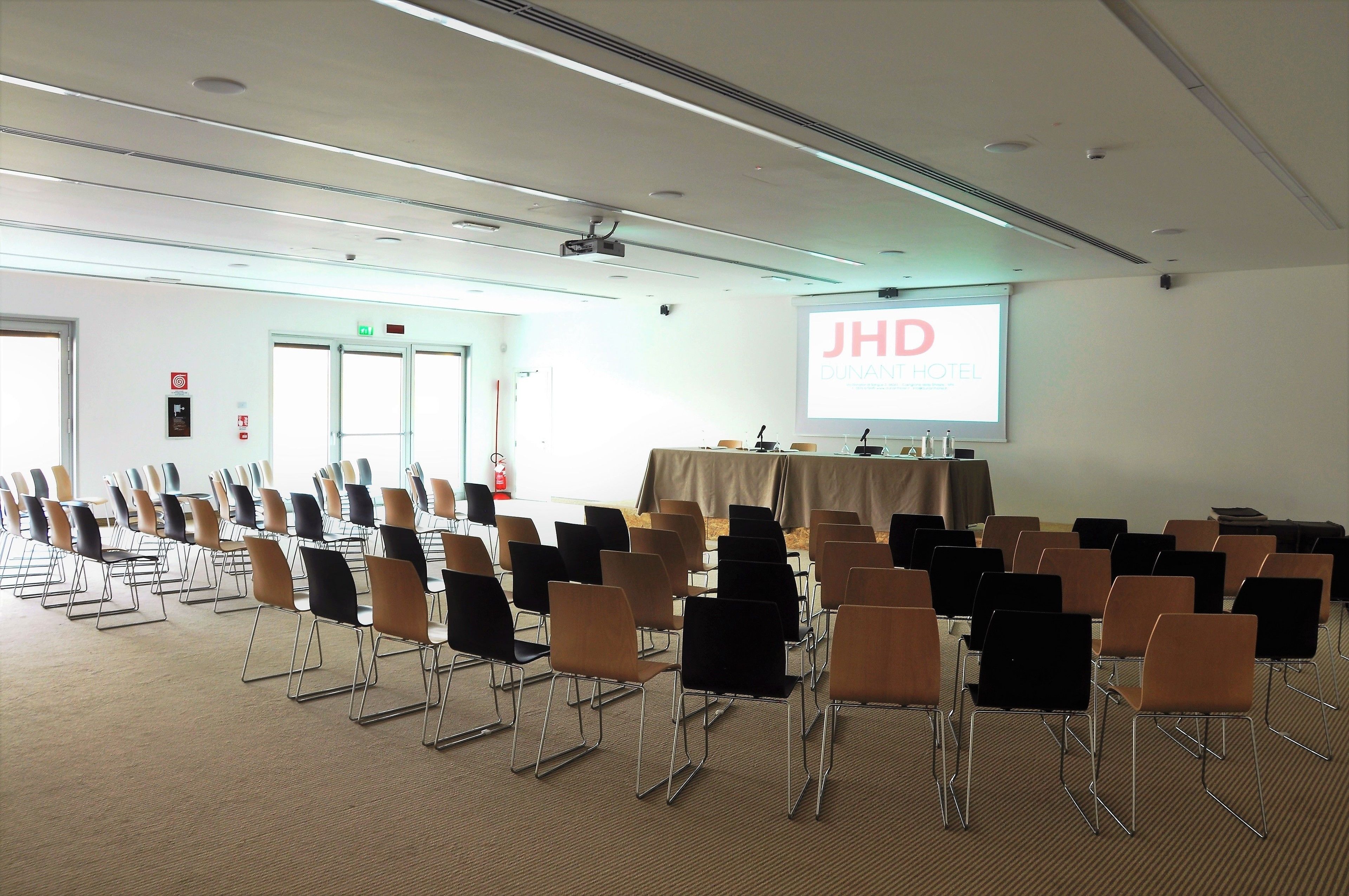 A conference room with rows of chairs, a projector screen displaying "JHD DUNANT HOTEL," and a table with a tablecloth.