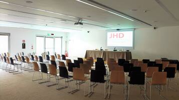 A conference room with rows of chairs, a projector screen displaying "JHD DUNANT HOTEL," and a table with a tablecloth.