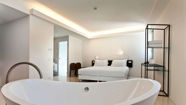 A modern bedroom with a large white bathtub, a bed with white linens, and a glass shelving unit. - Aiden by Best Western JHD Dunant Hotel (Castiglione delle Stiviere)