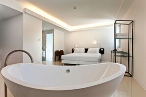A modern bedroom with a large white bathtub, a bed with white linens, and a glass shelving unit.