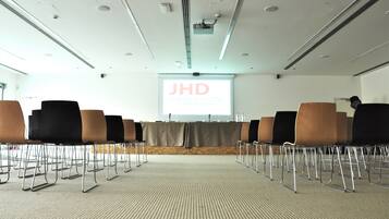 Ett konferensrum med rader av stolar, en projektionsduk som visar texten ”JHD DUNANT HOTEL” och ett golv täckt av matta.