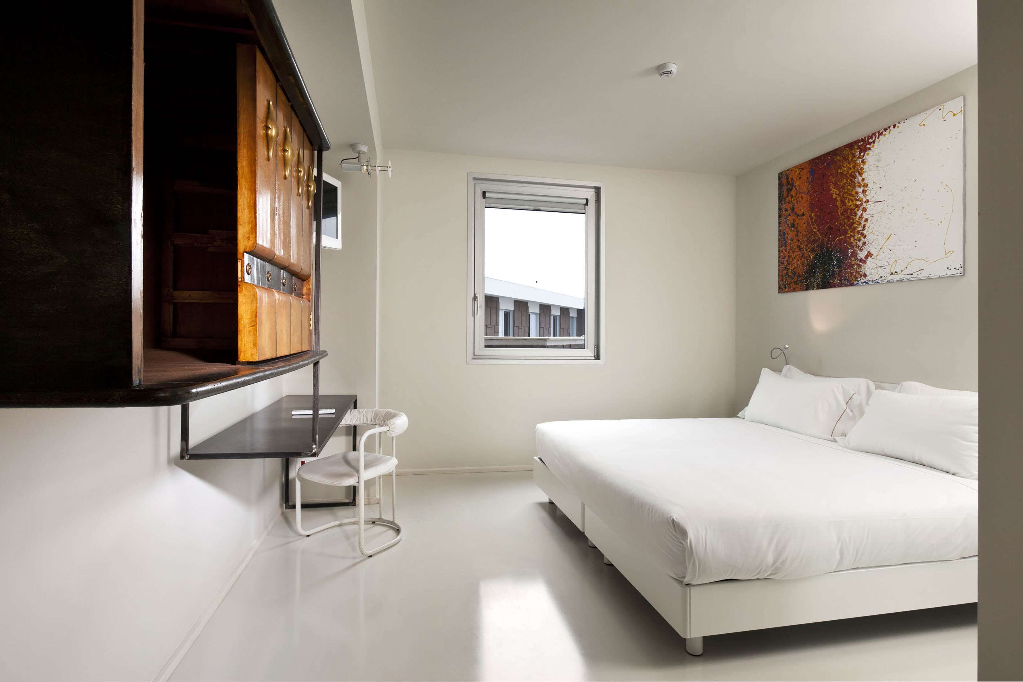 Un dormitorio moderno con una cama blanca, un escritorio pequeño, una silla, un ventanal y un cuadro en la pared.