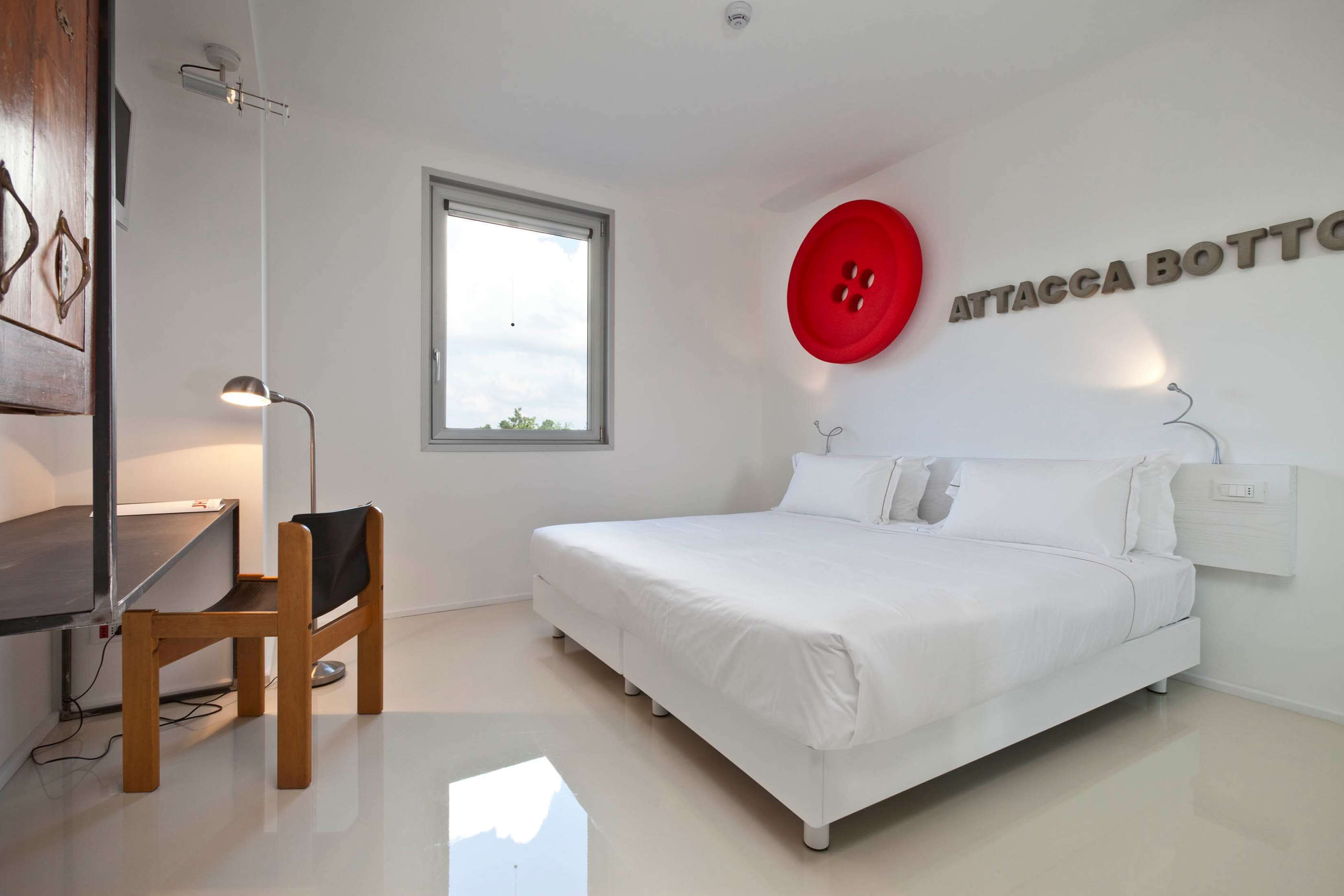 A modern bedroom with a bed, a desk, and a chair. There is a window and a wall-mounted red button.