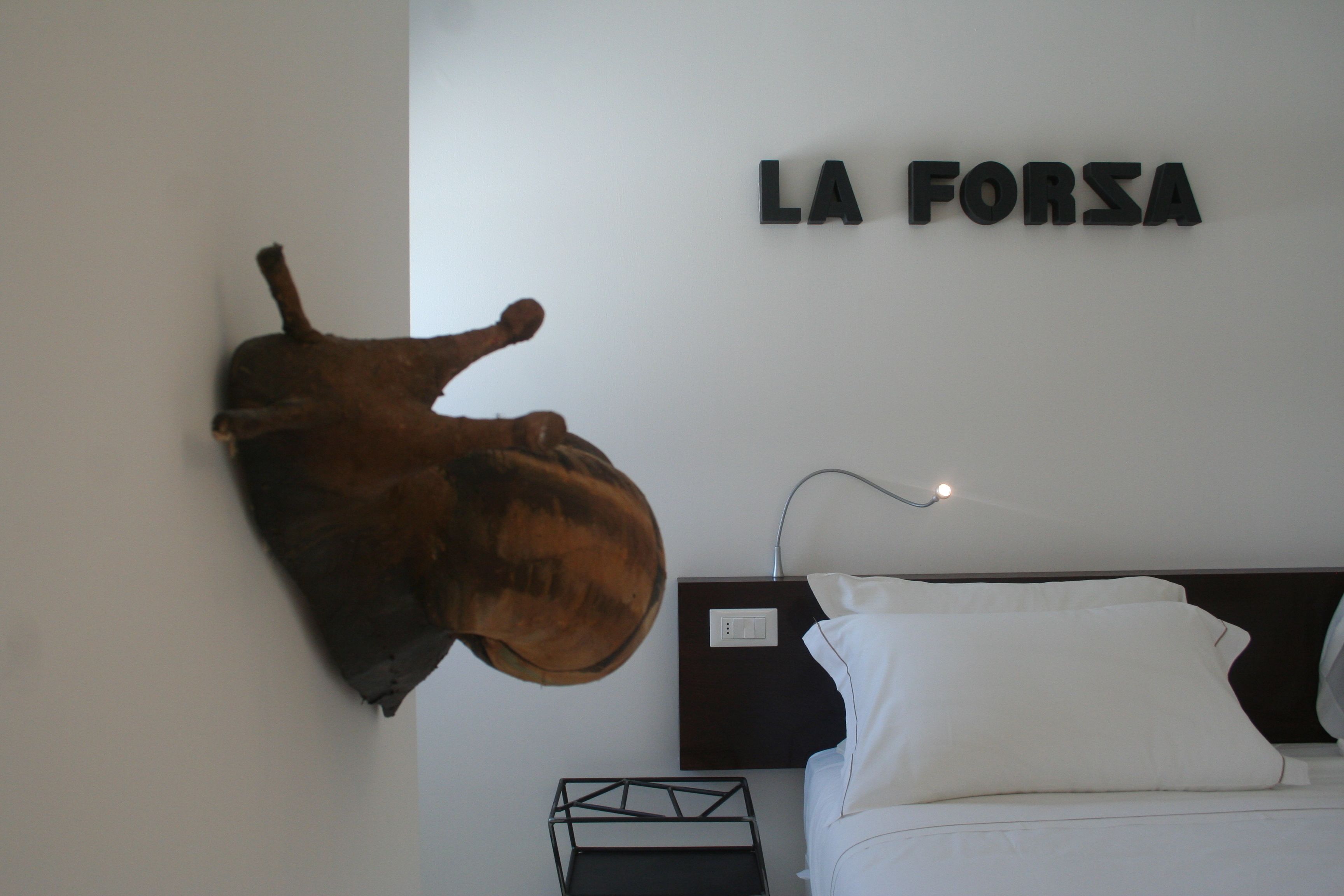 A room with a mounted sculpture, a bed with white bedding, and a wall-mounted sign reading "LA FORSA".