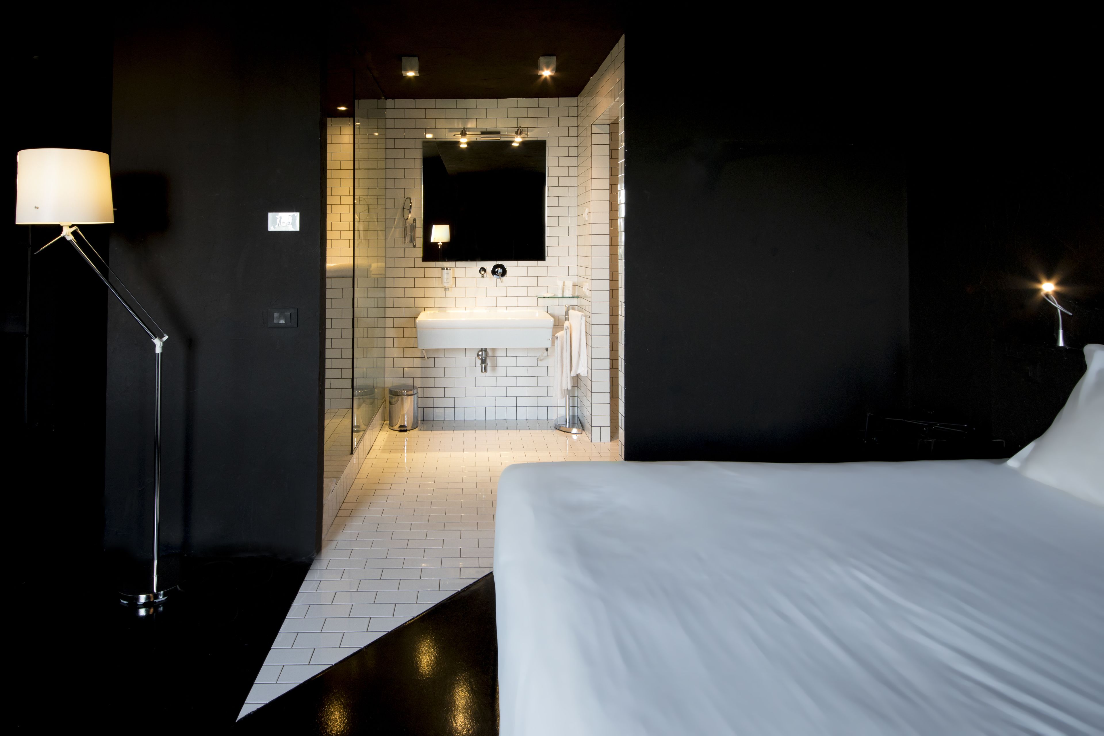 A hotel room with a large bed, a bedside lamp, a bathroom with a sink and mirror, and a dark wall.