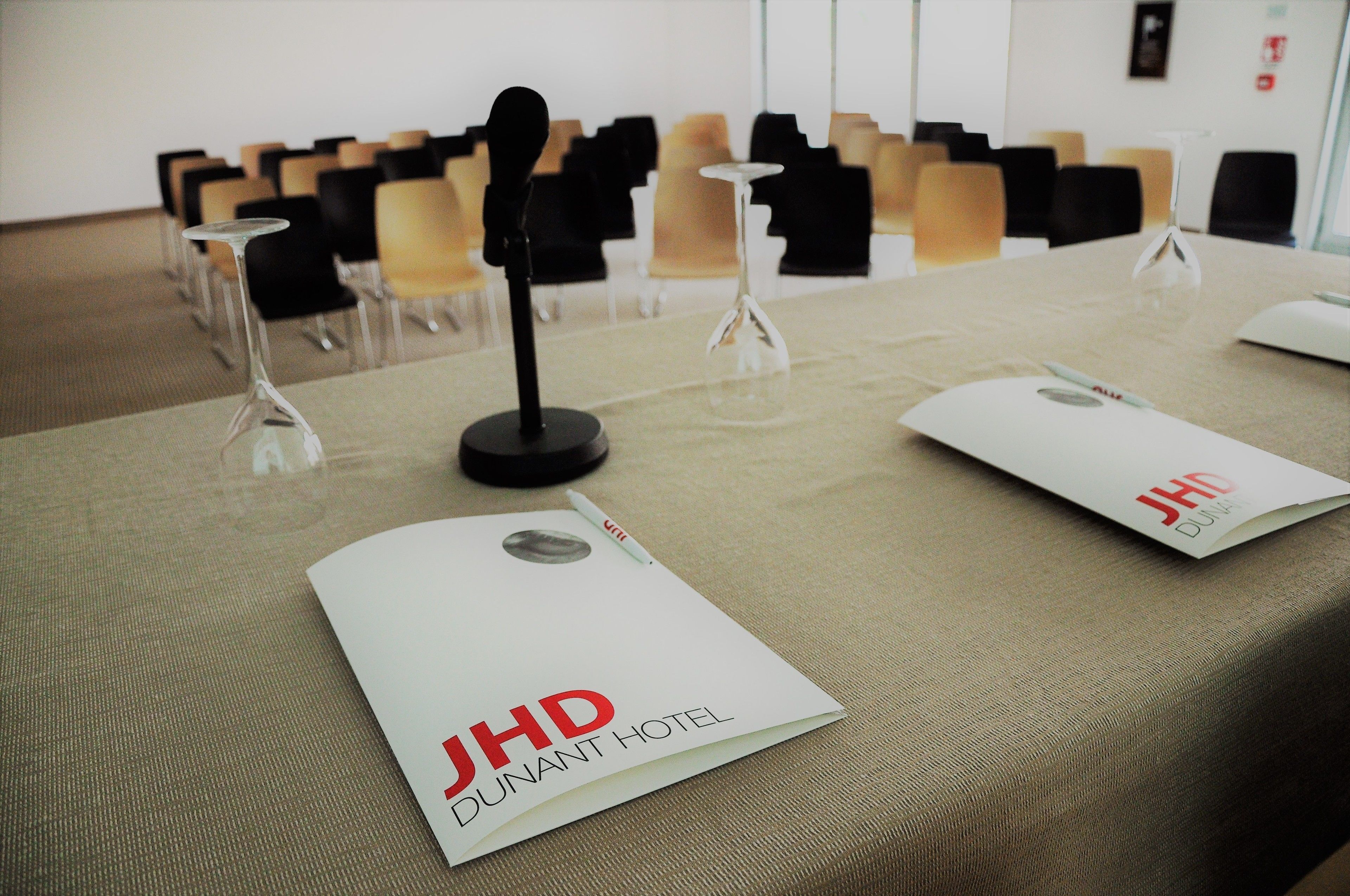 Una sala conferenze con un tavolo apparecchiato per una riunione, comprensivo di cartellini di nome, un microfono e una brochure per l'Hotel JHD Dunant.