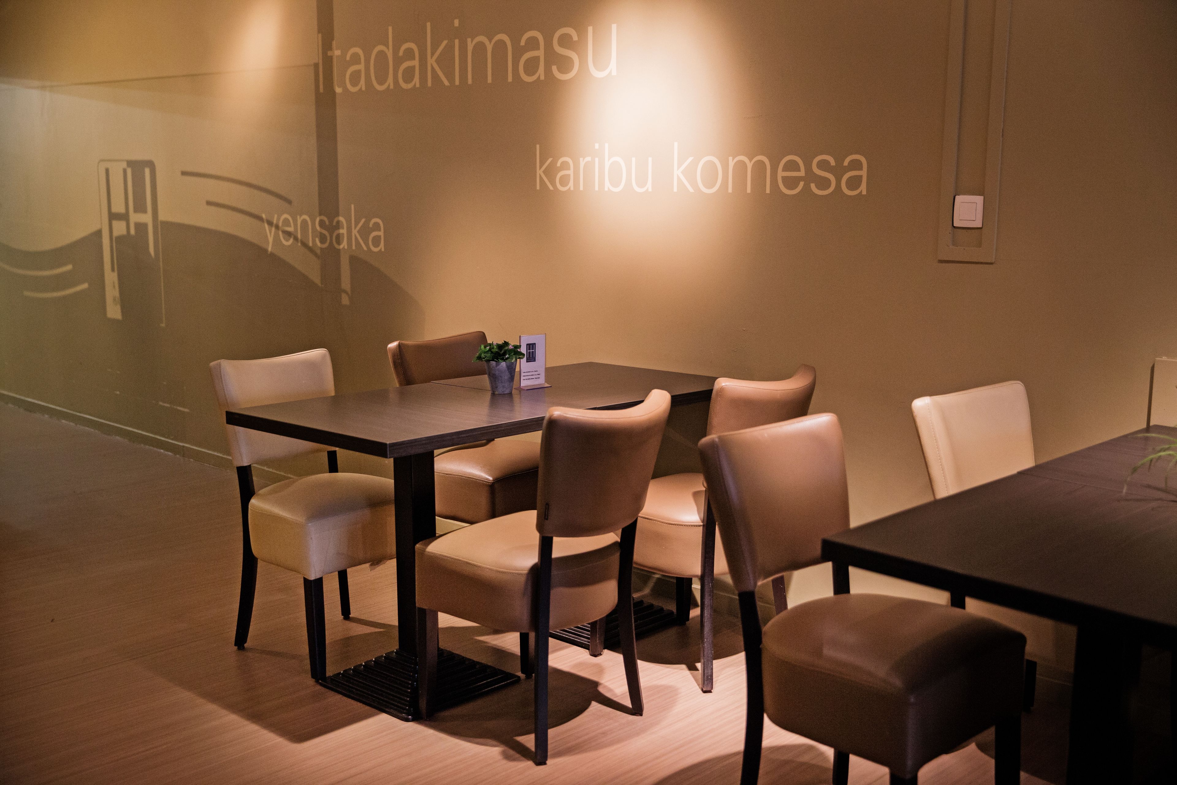 Een vergaderruimte met een donkere tafel en beige stoelen, een kamerplant en een projectiescherm met Japanse tekst.