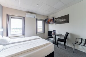 Quarto de hotel com cama, escrivaninha, duas cadeiras, televisão e janela com cortinas.