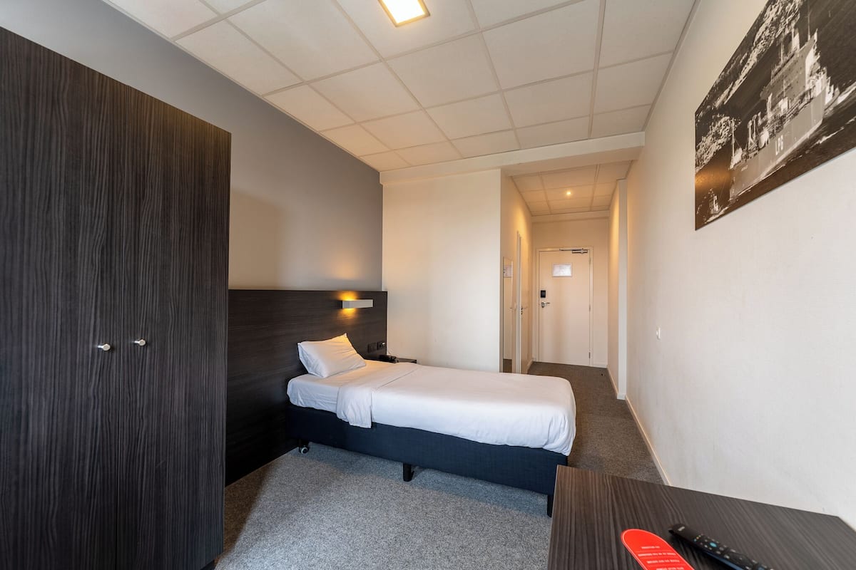 A hotel room with a single bed, a dark wooden wardrobe, a desk with a chair, and a small table with a remote control.