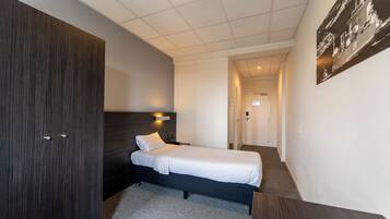 A hotel room with a single bed, a dark wooden wardrobe, a desk with a chair, and a small table with a remote control.