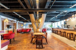 Un restaurante moderno con suelo de madera, sillas negras y rojas, y una mesa central con un jarrón de ramas.