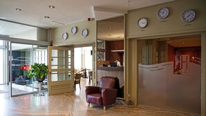 A lobby with a reception desk, a brown leather chair, and clocks displaying different time zones. - Antwerp Harbour Hotel (Antwerp)
