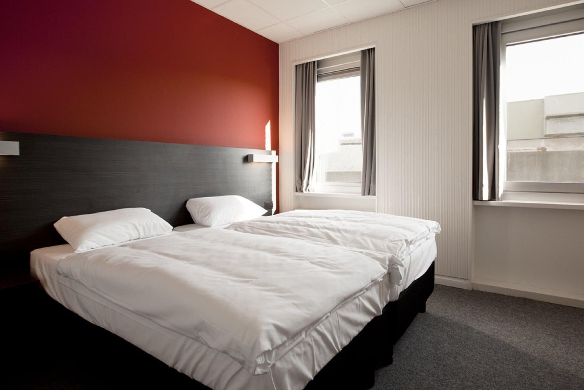A hotel room with a double bed, a headboard, a window with curtains, and a view of the cityscape.
