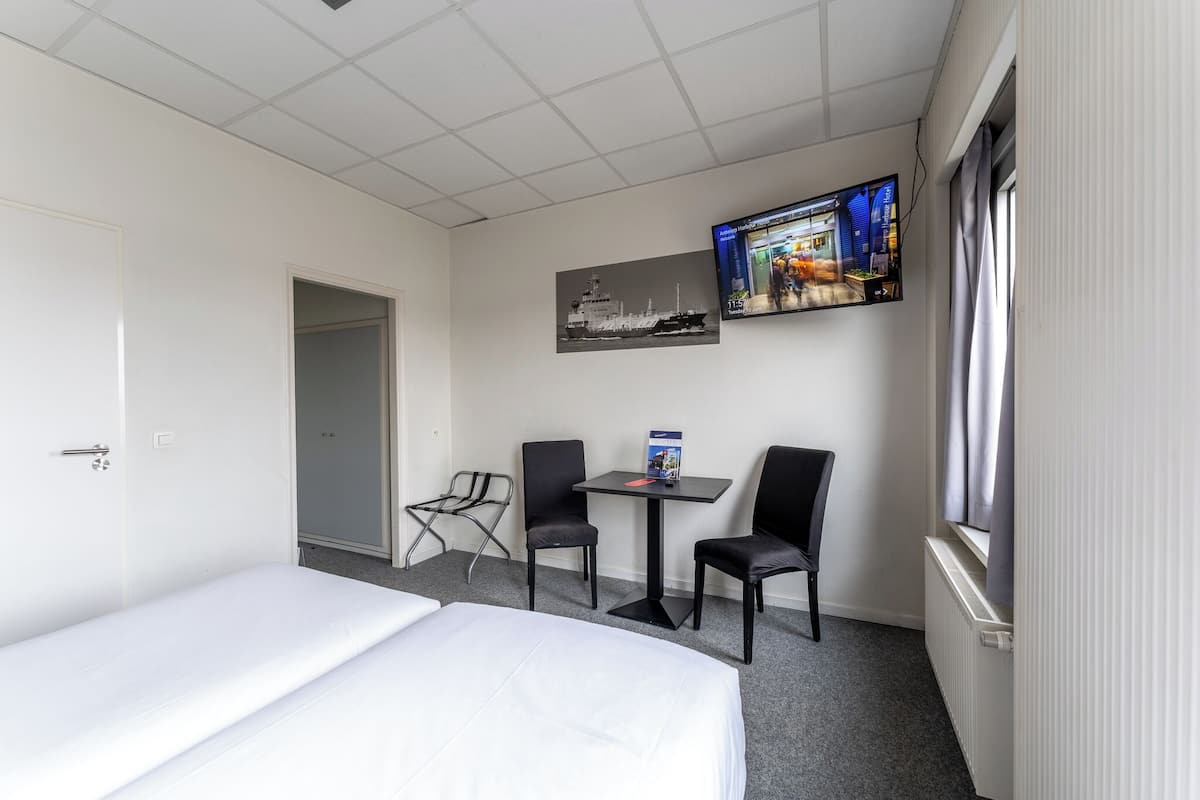 A hotel room with a bed, two chairs, a small table, a TV mounted on the wall, and a window with curtains.
