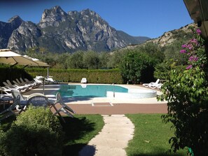 A pool area with lounge chairs and umbrellas, surrounded by greenery and mountains in the background. - Hotel Villa Rosa (Nago-Torbole)