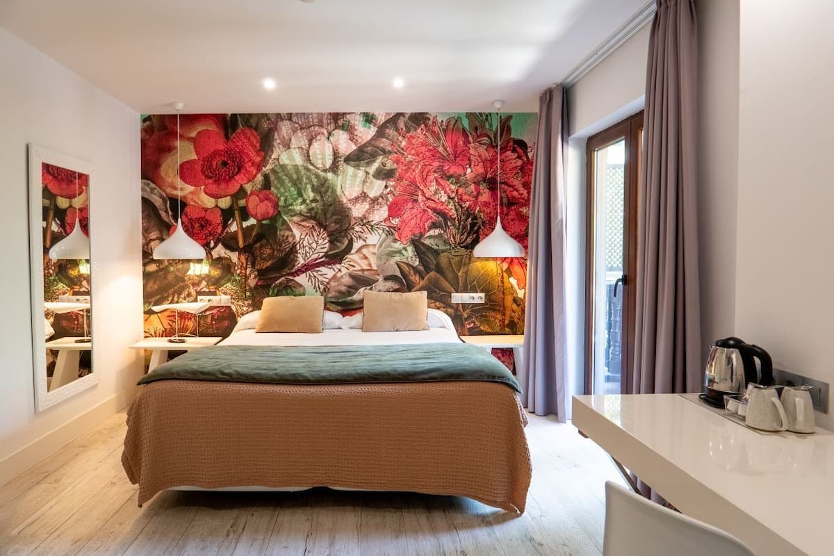 Habitación de hotel con un gran mural floral en la pared, una cama con cubrecama verde, una mesita de noche con lámpara y vistas de la ciudad a través de la ventana.
