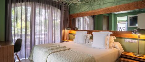 Un dormitorio con una cama grande, un escritorio y una silla. Hay una ventana con cortinas, un techo con diseño de hojas y un televisor montado en la pared.
