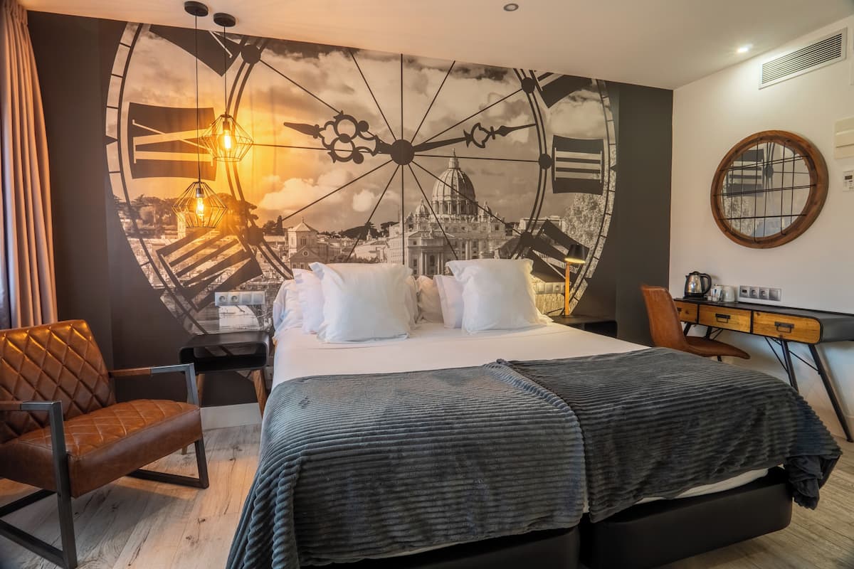 Habitación de hotel con un gran mural de un paisaje urbano, una cama con sábanas blancas, un sillón de cuero marrón, un escritorio de madera y un espejo redondo en la pared.