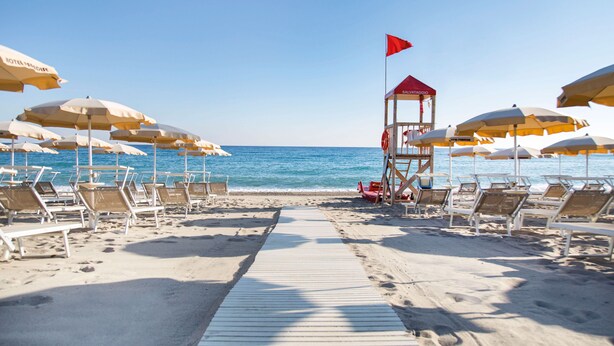 A beach with rows of sun umbrellas and lounge chairs, a lifeguard tower, and a clear view of the ocean. - Hotel Village Paradise (Mandatoriccio)