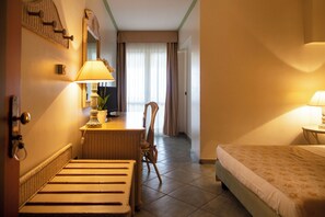 A hotel room with a bed, a desk with a lamp, a chair, and a view of the outside through a window with curtains. - Hotel Village Paradise (Mandatoriccio)