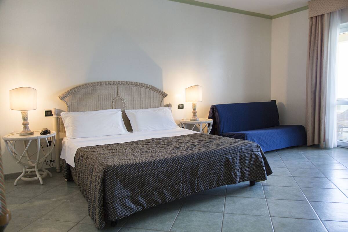 A hotel room with a bed, two bedside tables with lamps, a blue chair, and a window with curtains.