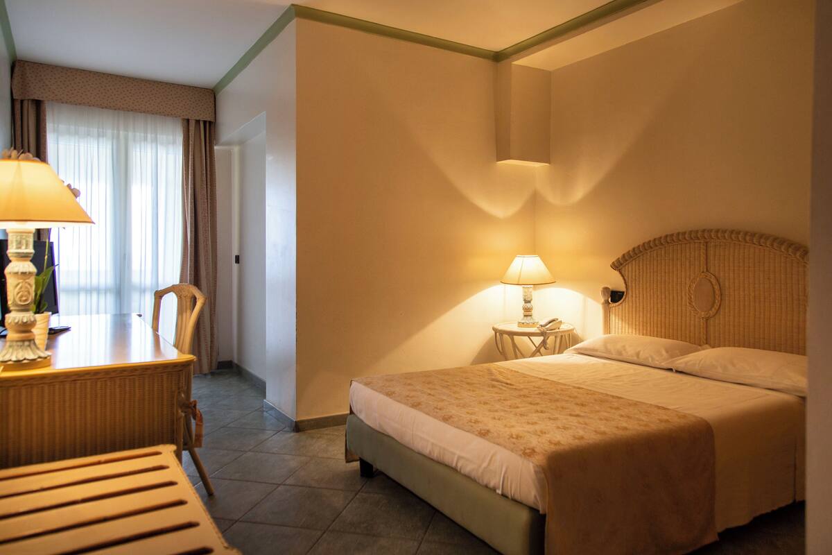A hotel room with a bed, two bedside lamps, a desk with a lamp, and a chair.