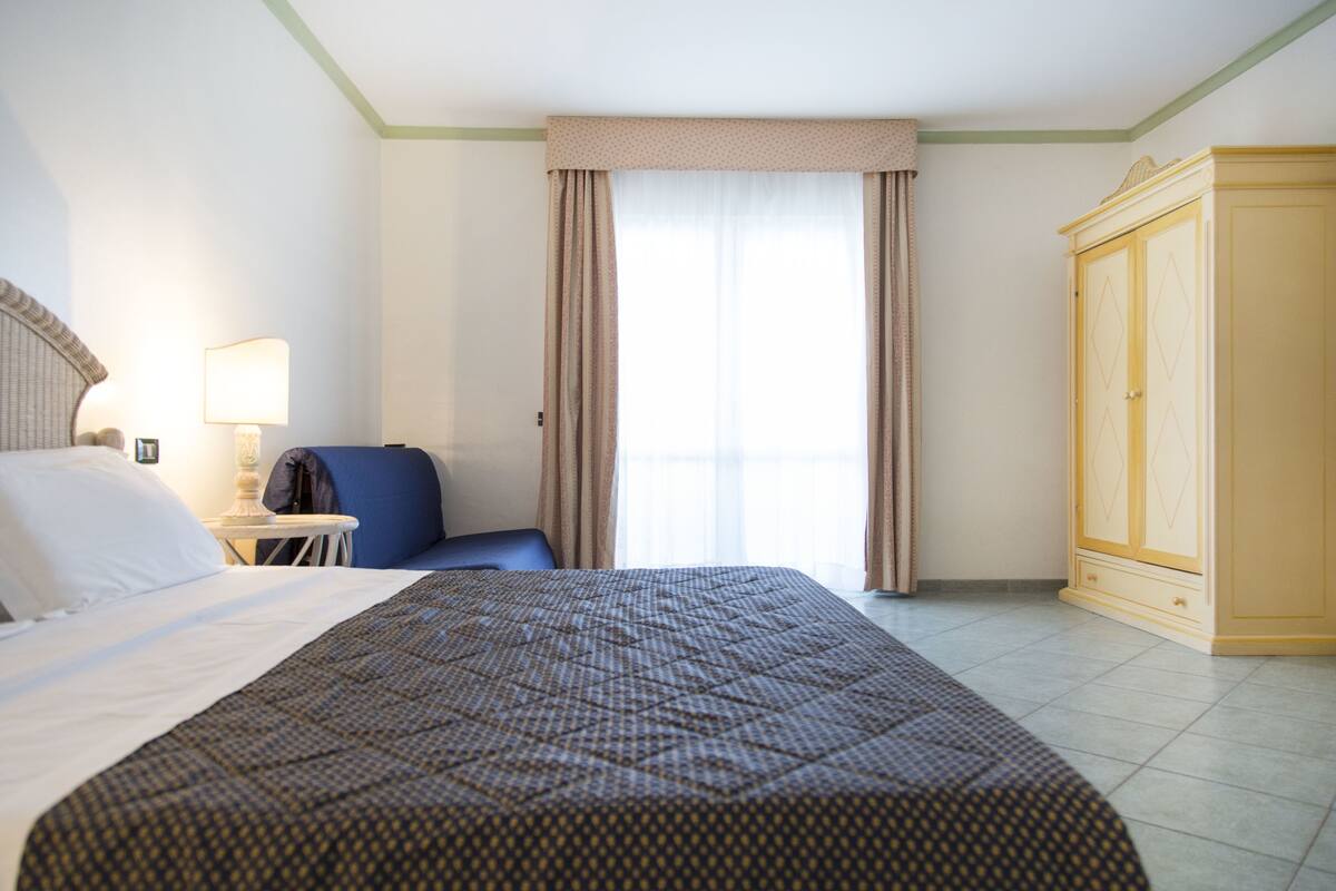 A hotel room with a bed, a chair, a wardrobe, and a window with curtains.