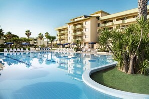 A pool area with lounge chairs and umbrellas, surrounded by palm trees and a multi-story building. - Hotel Village Paradise (Mandatoriccio)