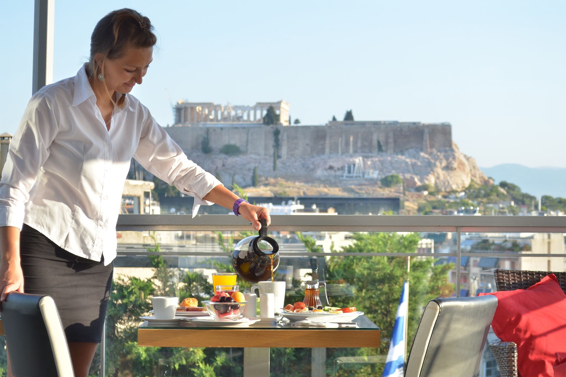 Una persona sirve café en una taza en un balcón con una mesa lista para el desayuno, con vistas a una fortaleza histórica.