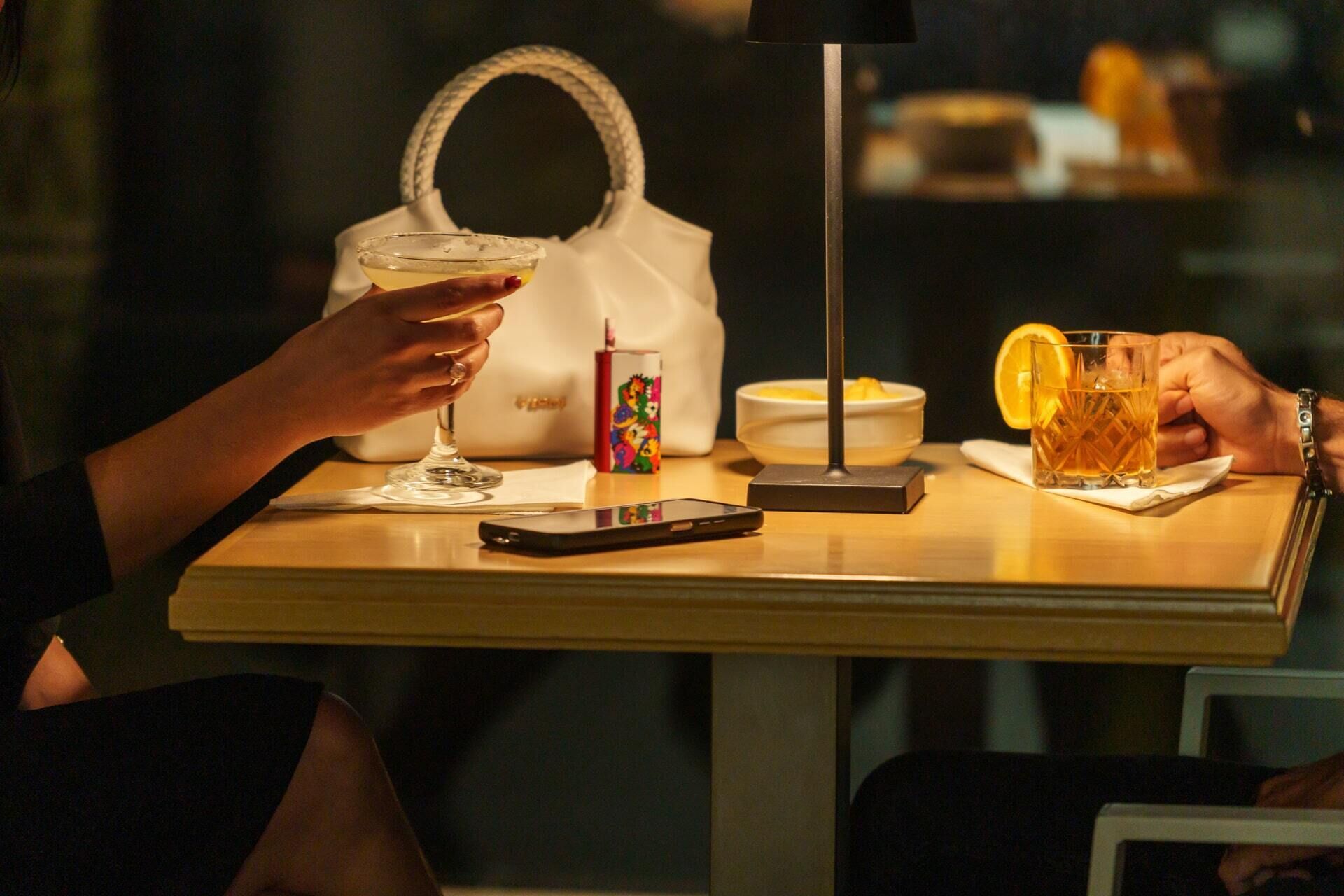 A table with a glass of amber liquid, a white handbag, a small bowl, and a smartphone.