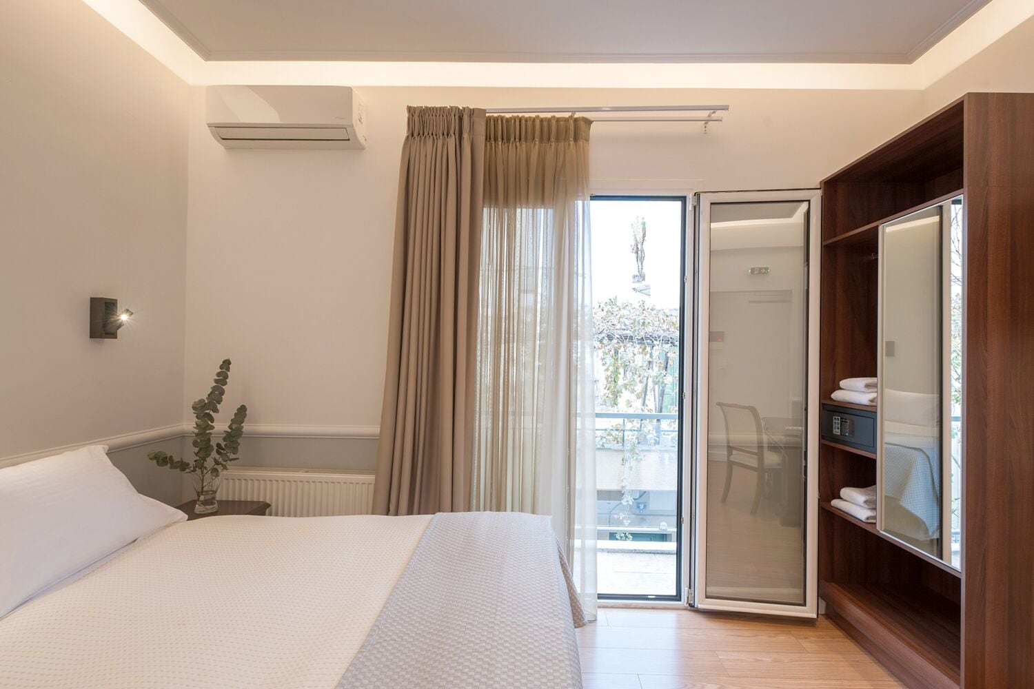 A hotel room with a bed, a wardrobe, a radiator, a potted plant, and a view of the outdoors.