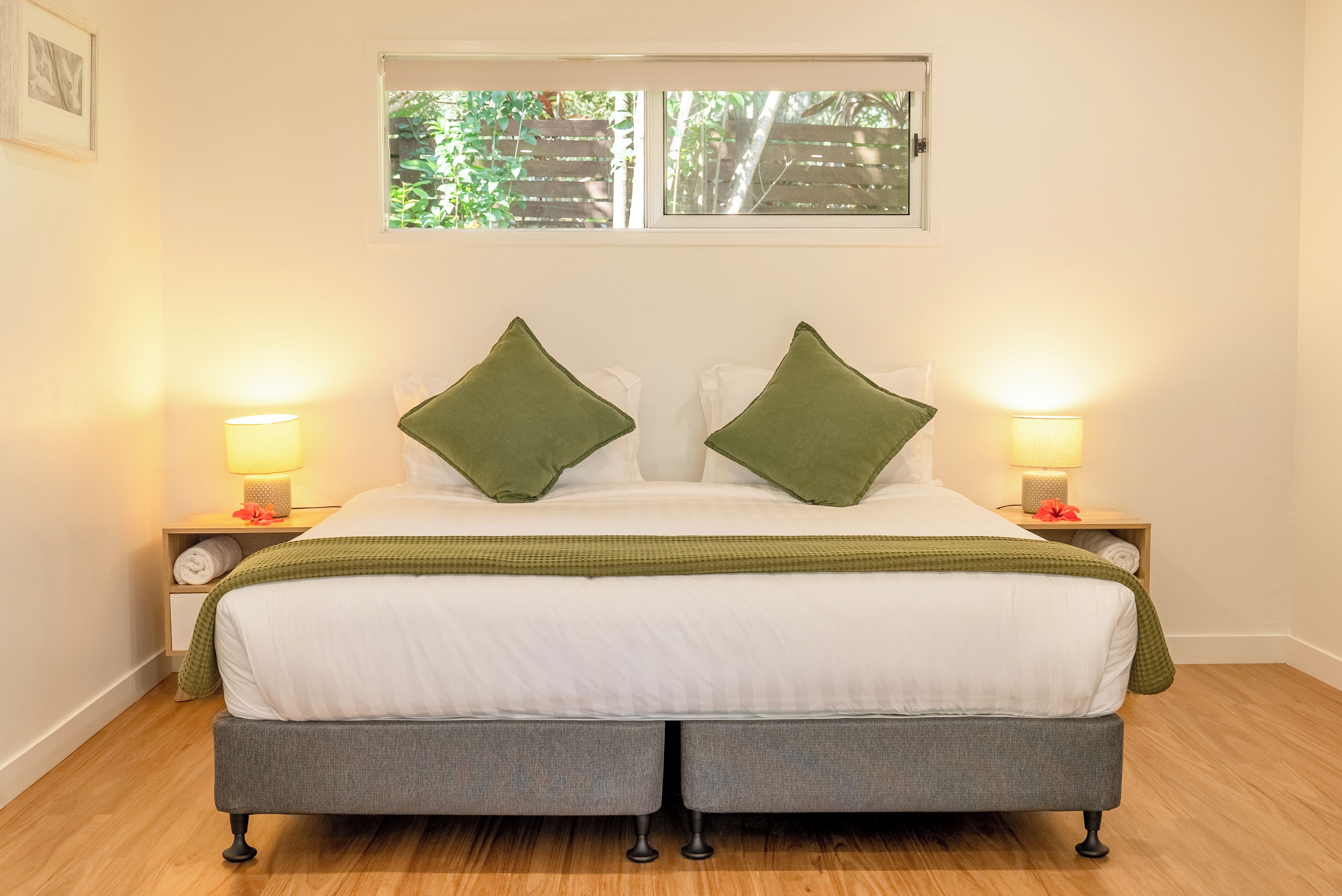 A neatly made bed with green pillows, two bedside lamps, and a window with a view of greenery.