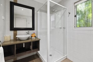 A modern bathroom with a white sink, a mirror, a glass shower enclosure, and a window with a view of greenery. - Broad Leaf Villas (Norfolk Island)