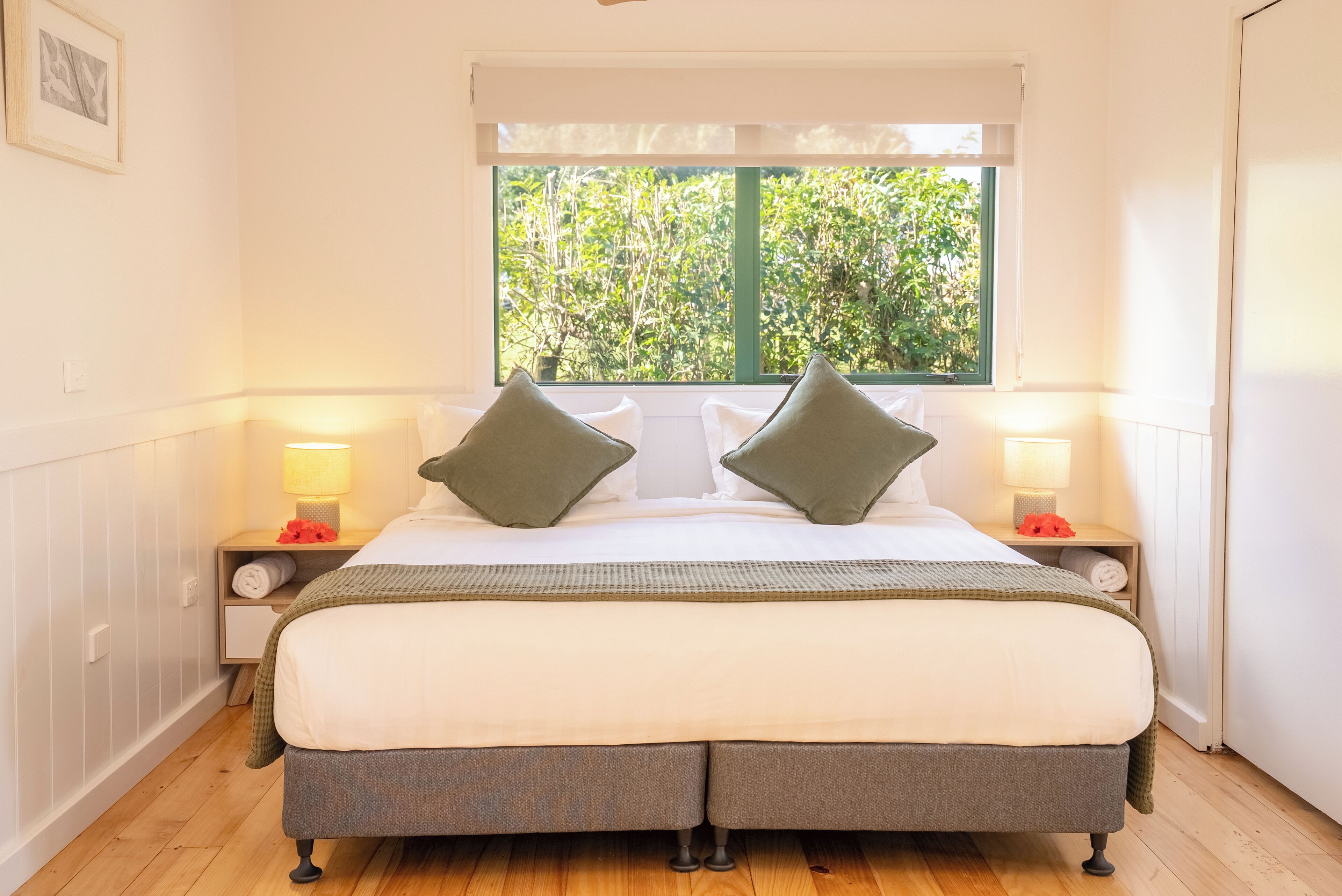 A double bed with two pillows, a bedside table with a lamp, and a window with a view of greenery.